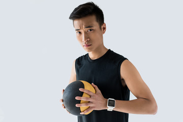 serious asian sportsman with smartwatch holding medicine ball isolated on grey background