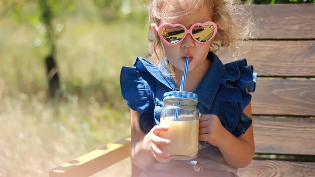 Child girl in sunglasses drinks smoothies in the park on a sunny windy day