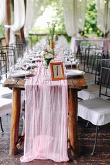 Wedding. Banquet. The festive table for guests, decorated with a composition of white and pink flowers and greenery, there are candles, served with crockery in wooden summer house.