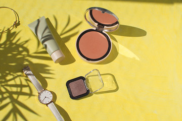 Female accessories Face powder Foundation Blush Watch Eyeshadows Bracelet Make up Summer holiday concept Flat lay Top view Copy space Tropical leaf shadow Yellow background