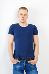 Young man in blue shirt and jeans poses with hands in pockets in white studio