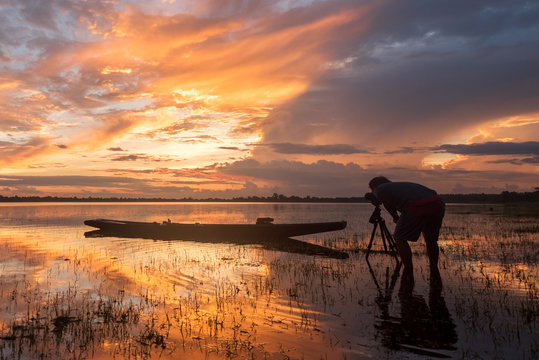 the photographer taking a picture of reflecion light of sunrise and fisherman boat at lake with vibrant sky , silhouette light