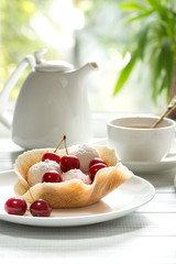 On a wooden white table are plate with  waffle basket with dessert, decorated with cherry berries, the  kettle and cup of tea