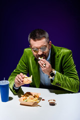 portrait of man in velvet jacket eating burger at table with french cries and soda drink with blue background