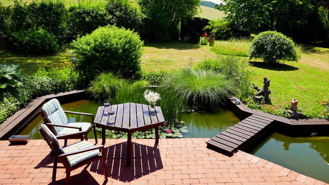 attraktiver naturbelassener Teich an rote Terrasse angrenzend mit Br&uuml;cke und Tisch und St&uuml;hlen