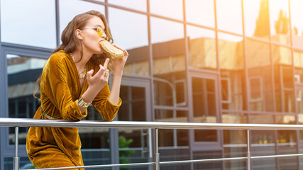woman in fashionable dress eating french hot dog and showing middle finger on street