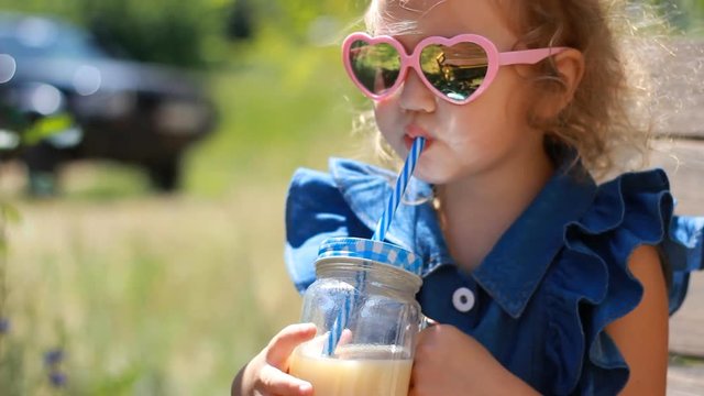 Child girl in sunglasses drinks smoothies in the park on a sunny windy day