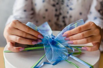 The hands of a young woman with a beautiful manicure tie a bow on a round gift box. Close-up, selective focus.