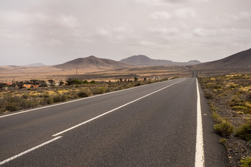 long way road from valley to mountains in the middle of nothing. fuerteventura vacation scenic place. arid and nobody location with grey sky and vulcans in background. asphalt lines and travel concept