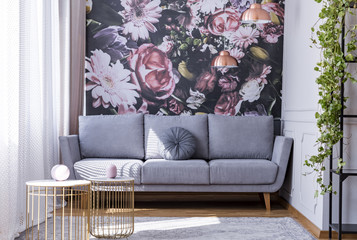 Real photo of bright sitting room interior with floral wallpaper, grey lounge with cushion, gold end tables and window with drapes