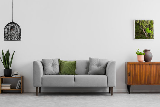 Grey sofa with pillows next to wooden cupboard in living room interior with lamp and poster. Real photo