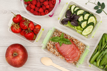 Apple and plastic meal prep containers with fresh strawberries, raspberries, boiled buckwheat porridge and slices of meat, cucumbers and salad, green peas