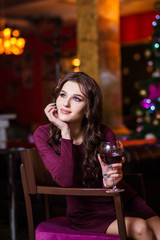 Christmas or New year celebration. Happy woman with a glass of champagne sitting near Christmas tree with xmas gifts. A fireplace with christmas stocking on background