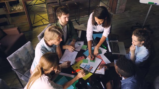 Multiracial workgroup is analyzing diagram and writing on stickers sitting at desk in office room. High angle view of people talking, pointing at charts and taking notes.
