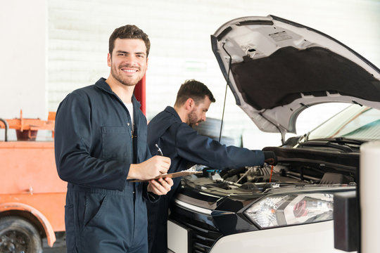 Technician Preparing Checklist While Colleague Inspecting Car In Garage