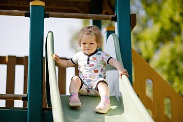 Happy blond little toddler girl having fun and sliding on outdoor playground. Positive funny baby child smiling.