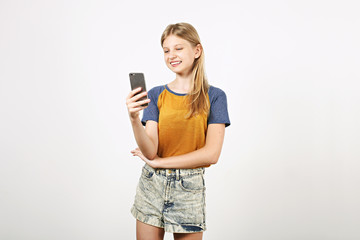 Portrait of young beautiful teenage girl with perfect smile laughing, holding mobile cell phone in hands. Happy young woman taking selfie with her smartphone, isolated on white background. Copy space.