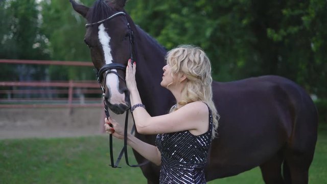 blond middle-aged woman 30-40. in an evening dress up to the floor hugs with a horse at sunset. Evening perfume makeup. attractive smart woman next to a brown horse 4K