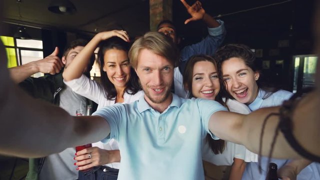 Group of colleagues are taking selfie together, young man is holding camera and posing, his coworkers are holding drinks in bottles, laughing and looking at camera.