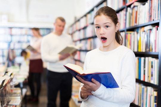 surprised tweenager girl standing in bookstore with book