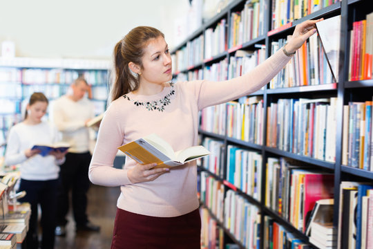 Young attractive female looking for necessary books on shelves in bookstore