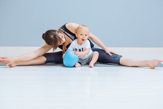 Happy Smiling Mother Stretching Legs. Mom Training Together With Sweet Positive Emtions Toddler Blond Baby Boy At Home Over Gray Wall Background. Fitness, Maternity And Healthy Lifestyle Concept.