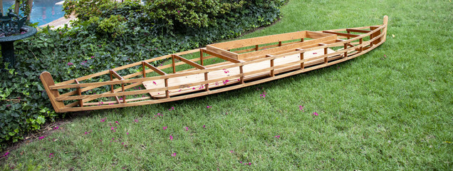 Building boats - a partially built wooden kayak sits on grass near a swimming pool and shubbery with pink flowers dropped from nearby crepe myrtle trees sprinkled around