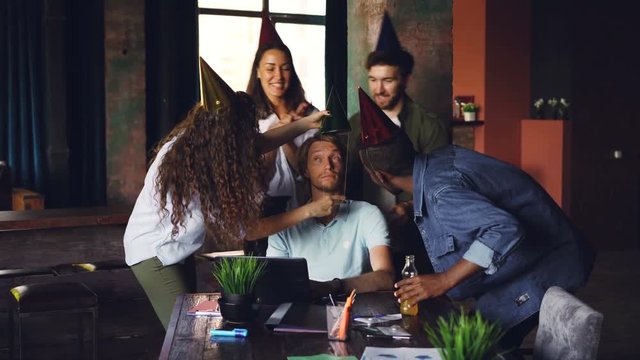 Company employees are congratulating their boss on birthday bringing cake and party hats, young man is blowing candles and doing high five while workers are clapping hands.