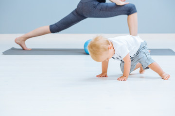 Naklejka premium Beautiful mother and little toddler son practicing yoga together at home, making stretching exercise, copy space. Kids crawling on foreground.
