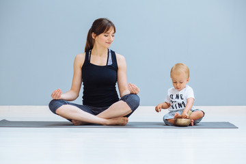 Fototapeta premium Cute european sporty mother with todller baby boy sitting together in lotus pose. Little son sitting with his mother on yoga mat over grey background. Motherhood, healthy lifestyle and people concept.