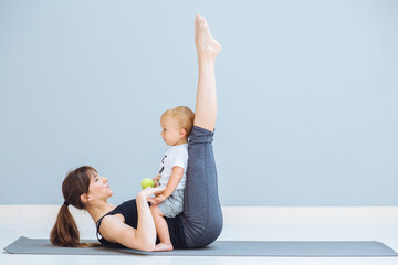 Sportive brunette mother with baby son doing press exercise on grey yoga mat over gray wall...