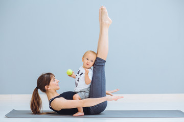 Sportive brunette mother with baby son doing press exercise on grey yoga mat over gray wall...