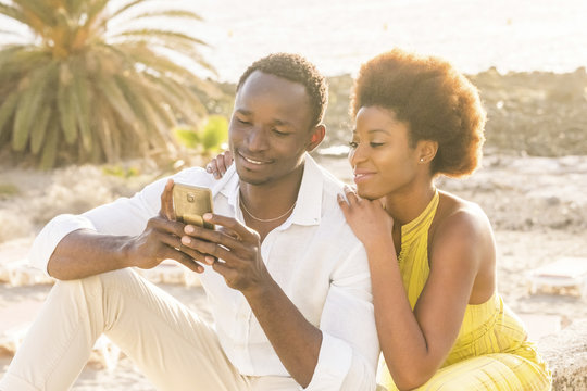 happy black young couple at the beach with sunset backlight on the background, smile and laugh using a smartphone to chat with friends or to see pictures of the vacation