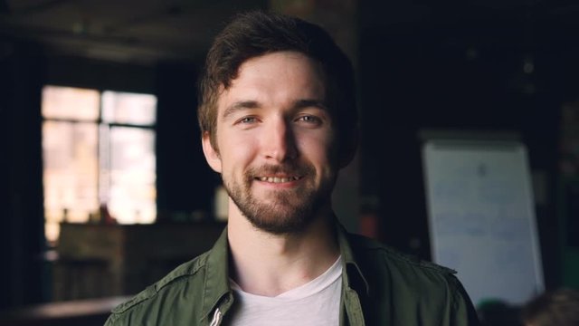 Close-up slow motion portrait of happy bearded guy with dark hair standing in modern office, looking at camera and smiling. People, work and career concept.
