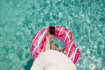 Fototapeta na wymiar top view of a woman relaxing in the pool with pink donuts in hot sunny day. Summer holiday idyllic. Enjoying suntan Woman in bikini and a hat. Holidays and summer lifestyle. She is using mobile phone