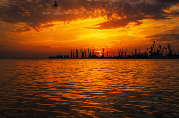 Beautiful landscape with fiery sunset sky and sea. Harbor on the coast during sunrise. Cranes silhouettes against fiery, orange and red sky.