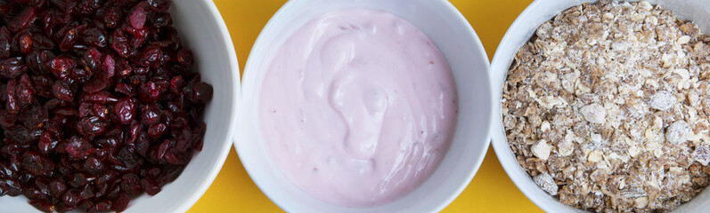 Three white bowls isolated, with yogurt cream, oat melas, crunch granola and driet fruits. Mix of variety cereals for daily breakfast. Yellow background. Banner.