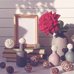 A fresh red rose with drops of water on the petals is in a gray vase. Nearby is a wooden frame, a stack of books, wicker balls and a variety of bottles. White wooden background