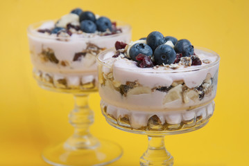 Breakfast or sweet dessert in glass cup. Two glasses of granola oat meals, with dried fruits, blueberries and yogurt. Isolated on yellow background.