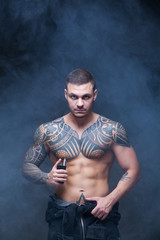 Vaper. The man with a muscular naked torso with tattoos smoke an electronic cigarette on the dark background