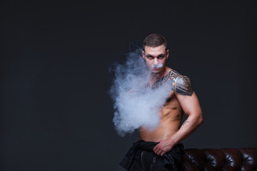 Obraz na płótnie Canvas Vaper. The man with a muscular naked torso with tattoos smoke an electronic cigarette on the dark background