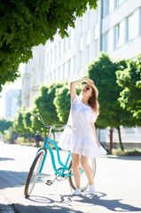 Female is standing on a city street looking up holding a vintage bicycle and a hat on her head. Lightness of movement, blond dress, sunglasses attached to the charm of photography
