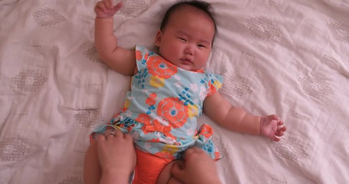 Asian mother is wiping off baby girl's tears and fixing her shirt while laying down on the white bed