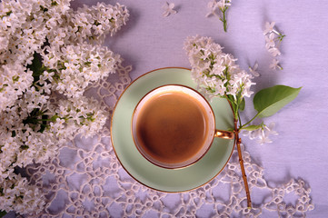 Closeup of flatlay arrangement of white lilacs and turquoise cup with coffee on tatted doily and white table cloth. Horizontal photo.