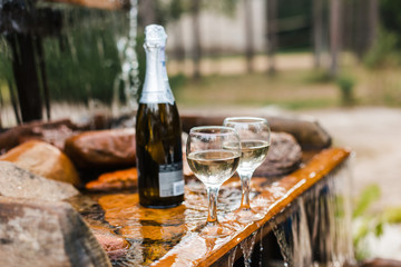 Love, romance, holiday, celebration concept. Bottle and glasses of champagne chilled by waterfall in summer forest on sunset.