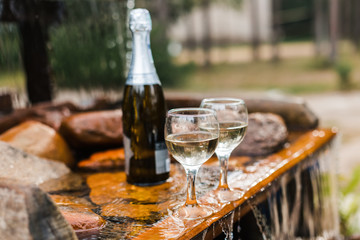 Love, romance, holiday, celebration concept. Bottle and glasses of champagne chilled by waterfall in summer forest on sunset.