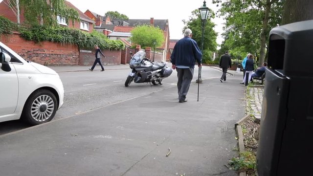 An elderly man walking down the pavement next to parked cars and a motorbike, using a walking stick.