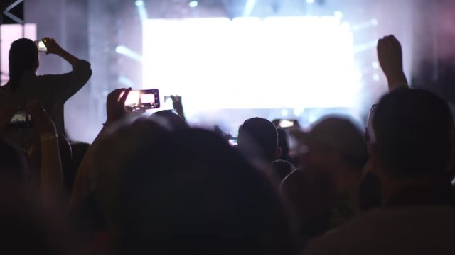 Crowd of people looking at the stage with big screen displaying unidentified rock musicians in bright lights of projectors