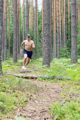 A young man with a muscular bare chest, in shorts and a cap, runs along a pine forest against the background of tree trunks