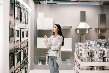 Half length portrait of brooding asian woman choosing new oven in furniture store